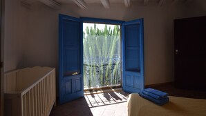 Ein Zimmer mit einem weißen Kinderbett, einer blauen Tür und Blick auf einen Kaktus durch ein vergittertes Fenster.