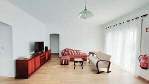 A living room with a sofa, a coffee table, a TV on a cabinet, and a hanging light fixture. - Family apartment in a quiet area 5 minutes from the beach! (Nazaré)