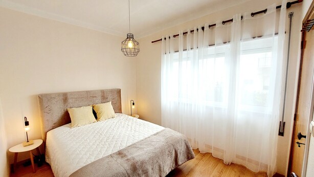 A bedroom with a large bed, a hanging light fixture, and sheer curtains. - Family apartment in a quiet area 5 minutes from the beach! (Nazaré)