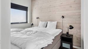 A bedroom with a wooden wall, a large window, a bed with white bedding, two bedside tables with lamps, and a nightstand with a lamp.