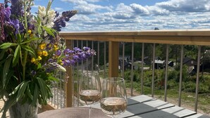 A dining table with a plate of food, two wine glasses, and a vase of flowers on a balcony with a view of houses and trees.