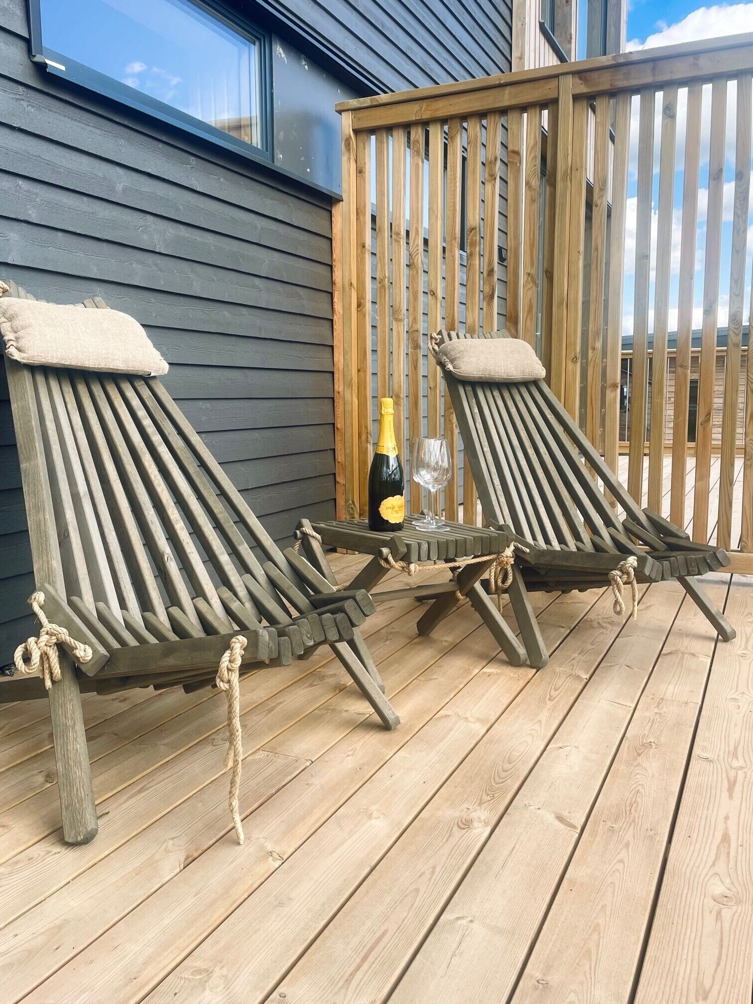 Two wooden lounge chairs on a wooden deck with a bottle of champagne and a glass.