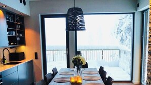 A modern dining area with a large table set for six, a hanging light fixture, and a view of a snowy landscape through large windows.