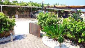 Un espace extérieur abrité, avec une voiture garée sous une pergola, entouré de différentes plantes et d’une terrasse en bois.