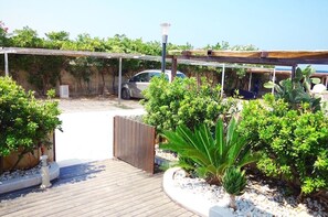 A covered outdoor area with a car parked under a pergola, surrounded by various plants and a wooden deck. - Seafront apartment with terrace, veranda & interesting extras (Villanova)