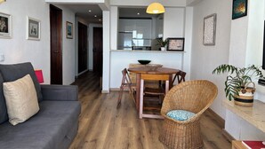 Un salon avec un canapé gris, un fauteuil en osier, une table à manger en bois et, au fond, une cuisine.