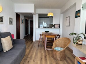 Un salon avec un canapé gris, un fauteuil en osier, une table à manger en bois et, au fond, une cuisine.