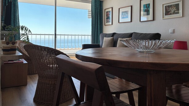 Une pièce avec une table à manger en bois, des chaises en osier, un canapé avec des coussins, une plante en pot et une vue sur l’océan par de grandes fenêtres.