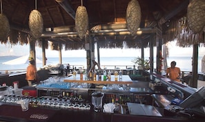 A bar area with a view of the ocean, bar equipment, and hanging light fixtures.