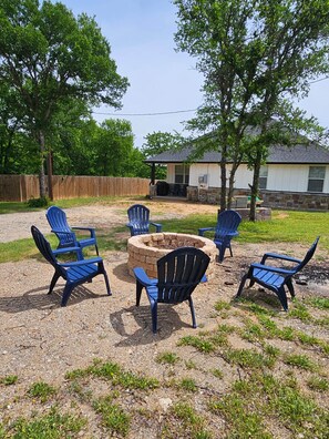 A backyard with a fire pit surrounded by blue Adirondack chairs, a wooden deck, and a house in the background.
