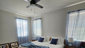 A bedroom with a bed, a ceiling fan, two windows with blinds, a nightstand, and a chair. - Maggie's Place (Granbury)