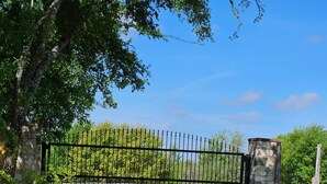 Une allée de gravier mène à une entrée fermée par un portail, avec un pilier en pierre et un portail en métal.