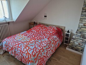 Ein Schlafzimmer mit schrägen Wänden, einem Bett mit gemustertem Überwurf, einem Nachttisch, einer Lampe und einer Ziegelwand.
