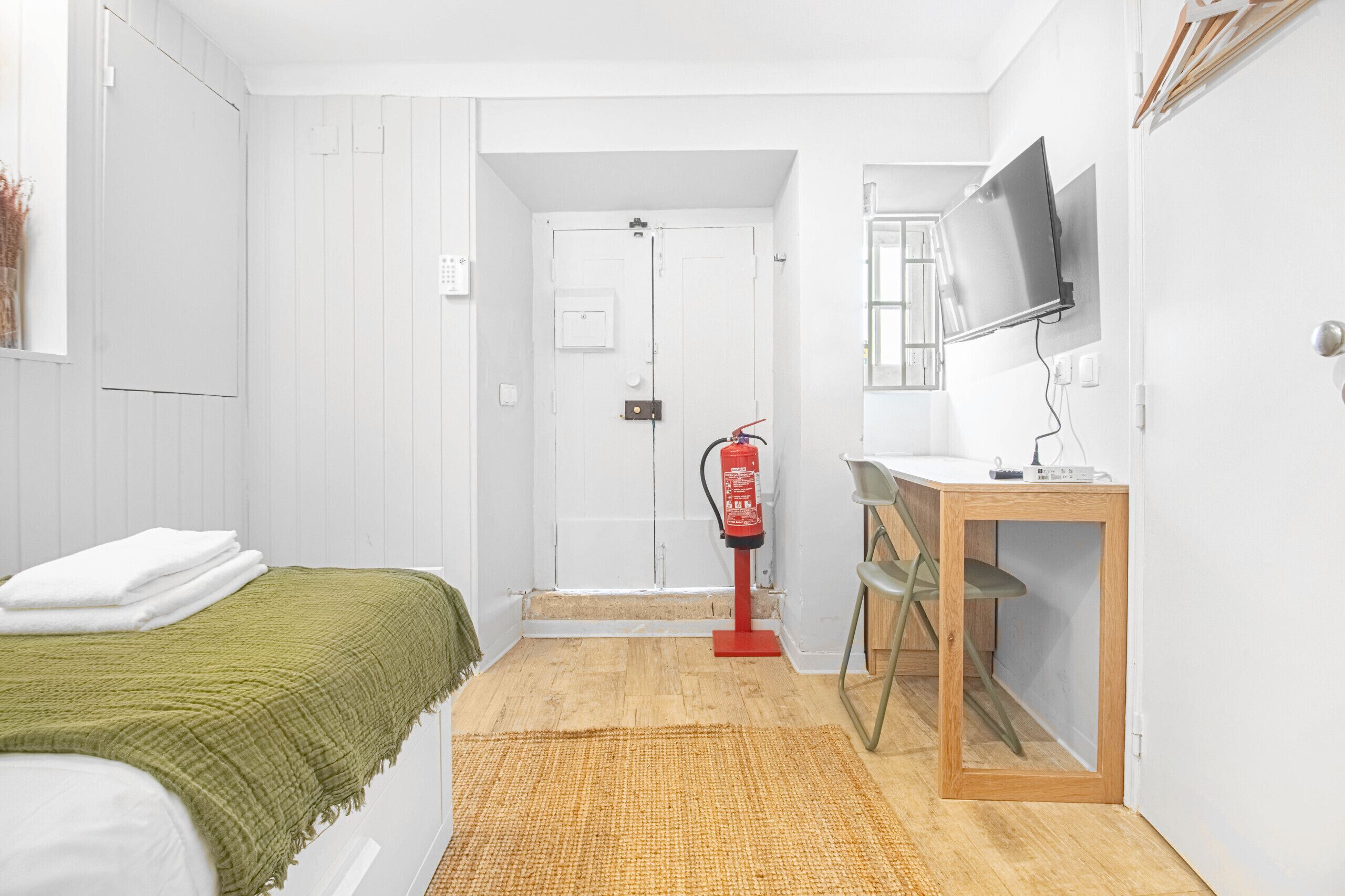 Una habitación con una cama, una mesita de noche con un espejo, un extintor y un televisor en la pared.
