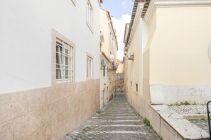 A narrow cobblestone alley between buildings with white and yellow facades, a stone wall on one side, and a metal railing on the other. - Nomad's Castel Lisbon - The Solo (Lisboa ciudad)