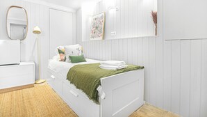 Un dormitorio blanco con una cama, una mesita de noche, una lámpara, un espejo y un cuadro colgado en la pared.