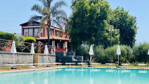 Une piscine avec des chaises longues et des parasols, un bâtiment à la façade rouge et un grand arbre en arrière-plan.