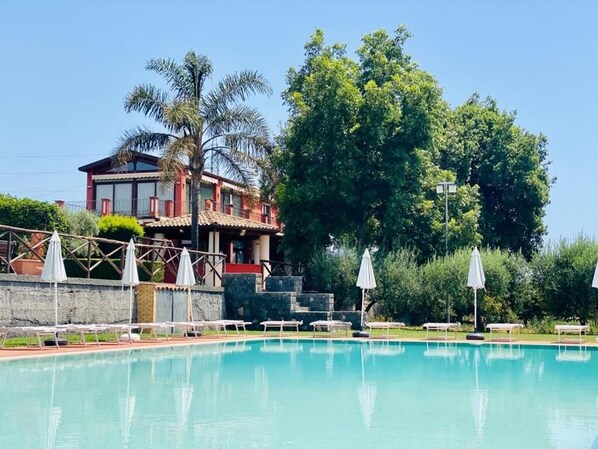 A swimming pool with lounge chairs and umbrellas, a building with a red facade, and a large tree in the background.
