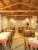 Un amplio comedor con vigas de madera en el techo, mesas dispuestas con manteles blancos y sillas, y un gran ventanal con cortinas.