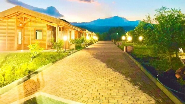 Un bâtiment en bois, accessible par un chemin pavé, entouré de verdure et avec une montagne en arrière-plan.