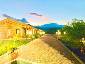 Un bâtiment en bois, accessible par un chemin pavé, entouré de verdure et avec une montagne en arrière-plan.