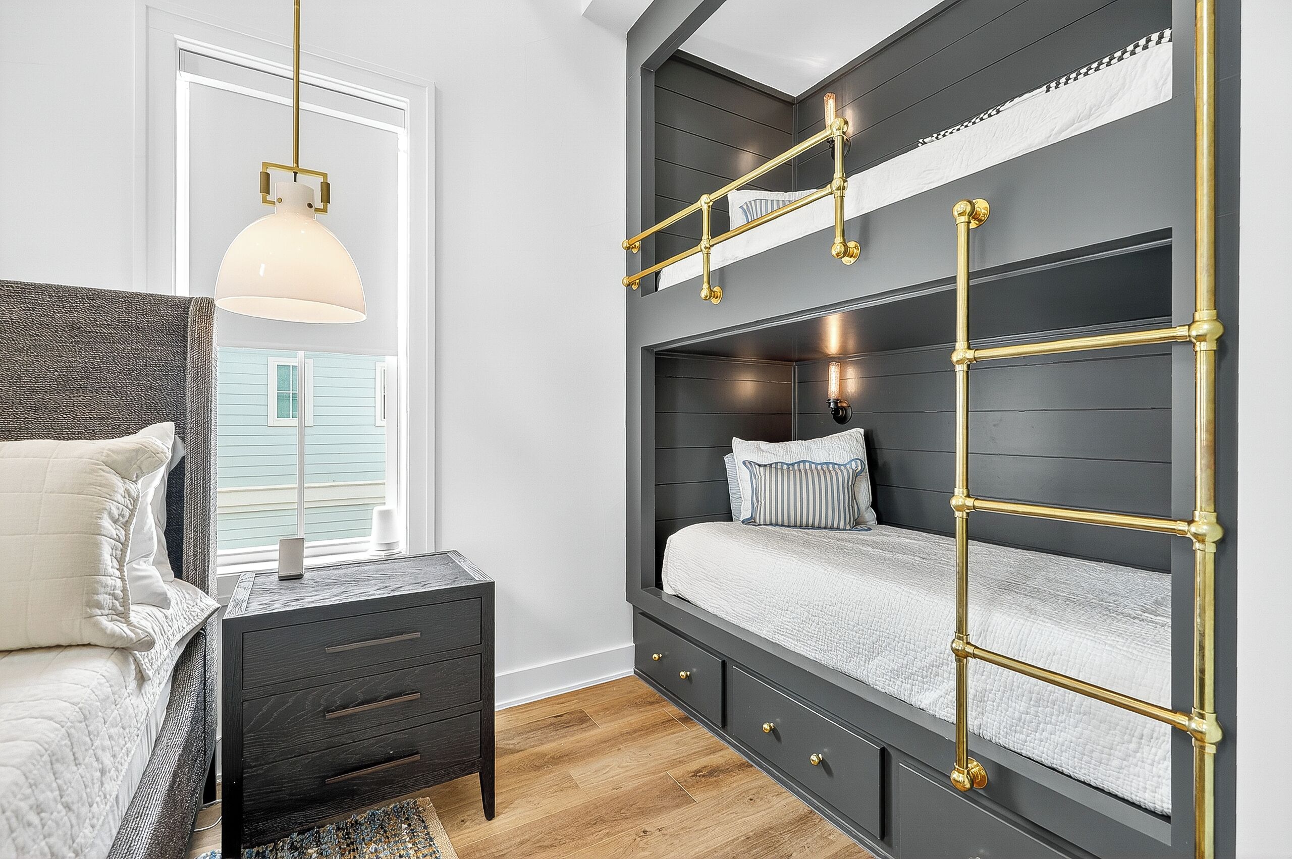 A modern bedroom with a bunk bed, a bedside table, a lamp, and a view of a house through a window.