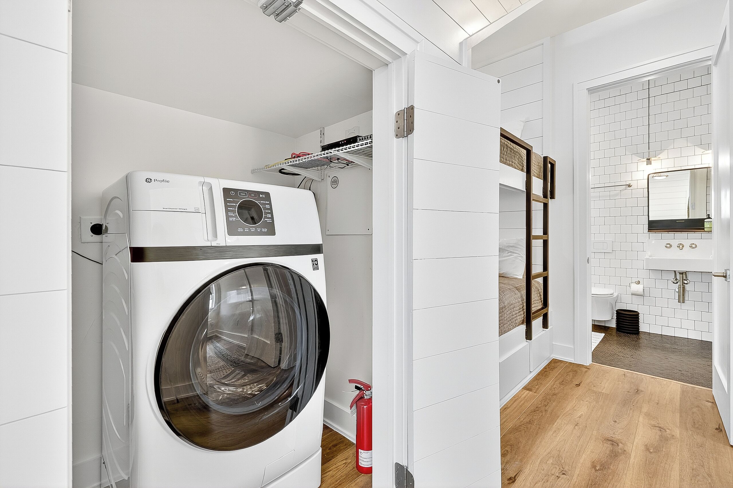 A modern laundry room with a front-loading washing machine, a fire extinguisher, and a small shelf with cleaning supplies.