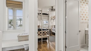 Ein kleines Zimmer mit Fenster, einem Bett mit kariertem Überwurf, einem Etagenbett, einem Deckenventilator und einer Tür mit gemustertem Wandpapier.