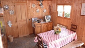 Ein gemütliches Blockhaushaus-Interieur mit Holzausbau, Steinböden, einem Tisch mit kariertem Tischtuch, einer hölzernen Bank, einem Fernseher auf einem Schrank und einem Fenster mit Blick ins Grüne.