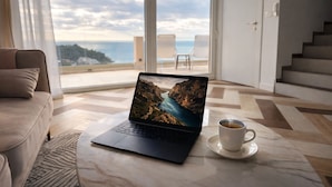 Ein Laptop auf einem Marmortisch mit Blick auf eine malerische Küstenlandschaft, eine weiße Tasse auf einem Untertasse und ein modernes Interieur mit Sofa und Treppe.