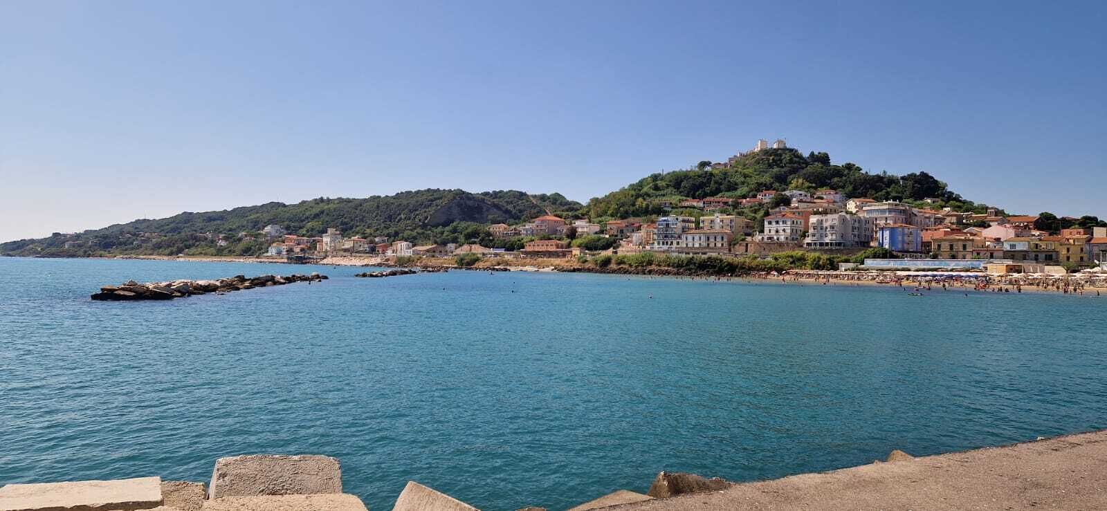 Un pueblo costero con edificios en una ladera, un mar azul cristalino y un rompeolas rocoso.