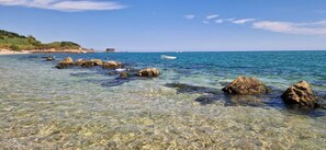 Un paysage côtier avec une eau peu profonde et claire, des rochers, un bateau au loin et une construction sur la plage.