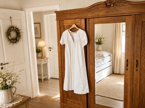 A bedroom with a wooden wardrobe, a hanging white dress, a bedside table with a lamp, and a bed with a floral arrangement. - Landhaus am See - Apartment Lärenberg, directly on Lake Edersee (Waldeck)