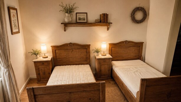 Una habitación con dos camas de madera, una mesita de noche con lámpara, un estante con libros y un florero, y una guirnalda colgada en la pared.