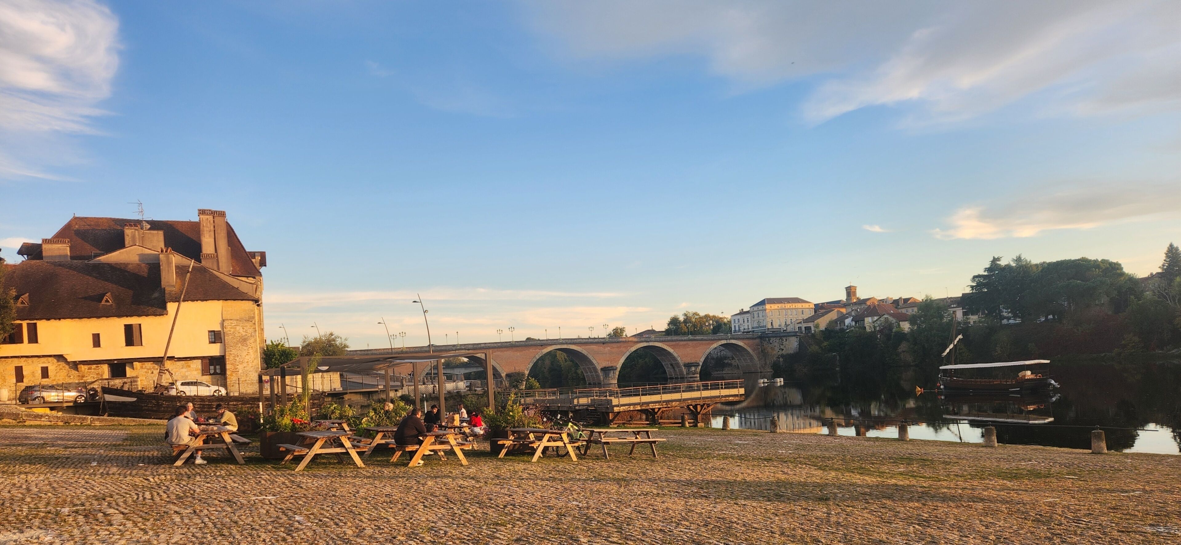Un área de picnic junto al río con bancos y mesas, un puente y edificios al fondo.