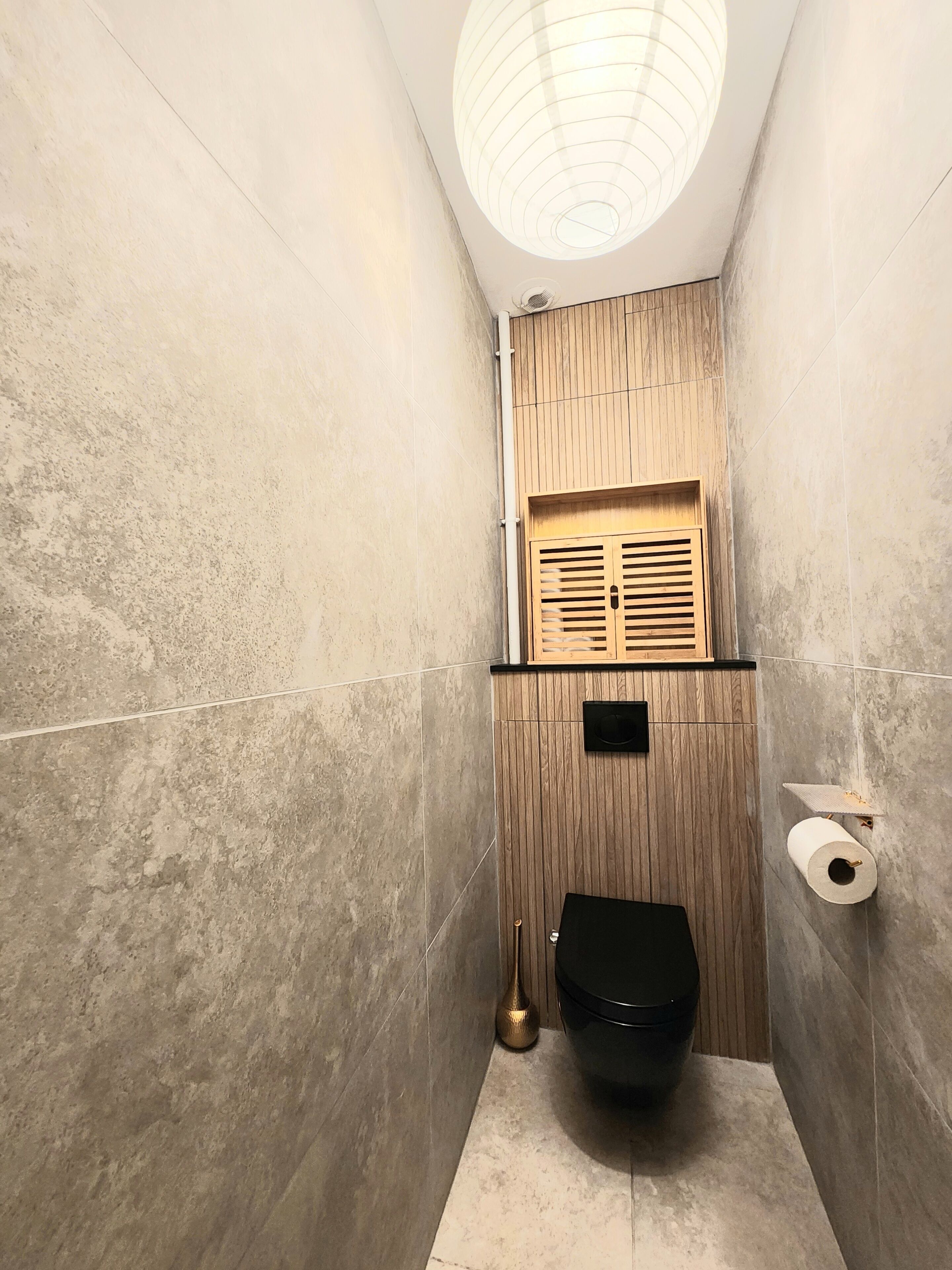 Un baño pequeño y moderno con inodoro, pared de paneles de madera, una lámpara en el techo y un perchero.