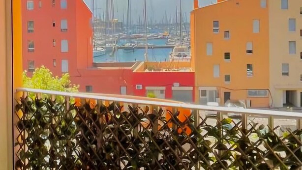 A view from a balcony with a metal railing, overlooking a marina with several sailboats and buildings with red and orange facades. - Apartment in cosy residence with magnificent view of the port (Occitanie)
