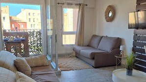 A living room with a sofa, a coffee table, and a TV. There is a balcony with a view of buildings. - Apartment in cosy residence with magnificent view of the port (Occitanie)