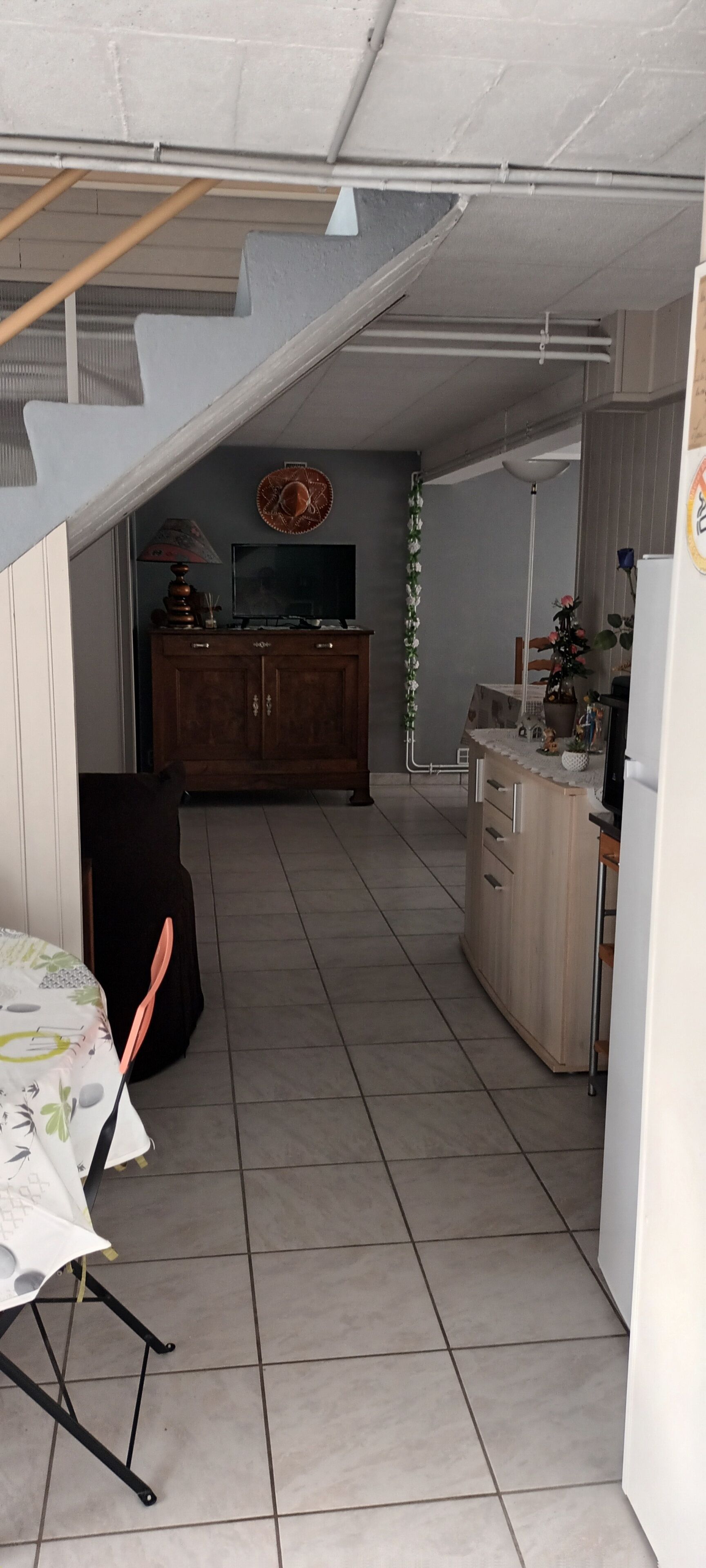 A narrow hallway with a dining table and chairs on the left, a wooden cabinet with a television in the center, and a refrigerator on the right.