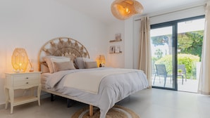 Un dormitorio con cama, mesitas de noche, un tapete redondo tejido y vistas al exterior a través de un ventanal corredero.