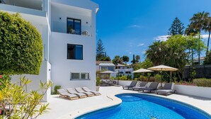 Un área de piscina moderna con sillones reclinables y sombrillas, rodeada de exuberante vegetación y un cielo azul brillante.
