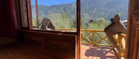 A room with a wooden floor, a large window with a view of mountains, and a wooden railing outside.