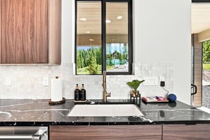 Una cocina moderna con encimera de mármol, un ventanal con vistas a un bosque y un lago, y un armario de madera.