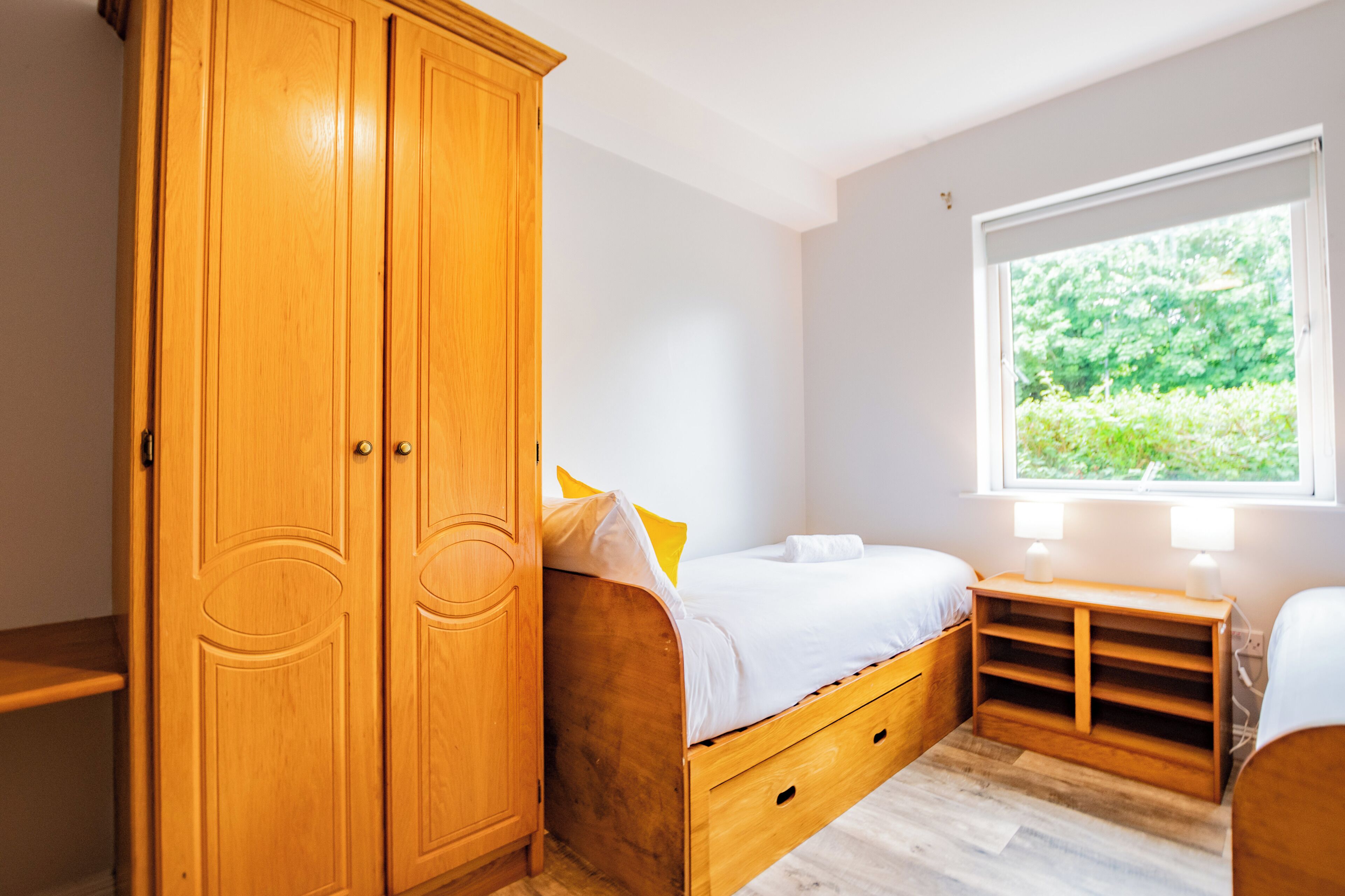 Ein Schlafzimmer mit Holzschrank, einem Bett mit weißer Bettwäsche, einem Nachttisch mit Lampen und einem Fenster mit Blick ins Grüne.