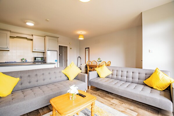 A living room with two grey sofas, a wooden coffee table, and a kitchen area in the background.