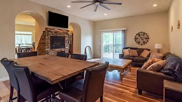 A spacious living area with a wooden dining table, dark chairs, a stone fireplace, a ceiling fan, and a large window.