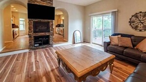 A living room with a stone fireplace, wooden flooring, a flat-screen TV, a large window, and a clock on the wall.