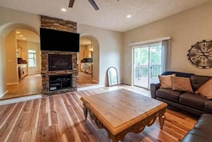 A living room with a stone fireplace, wooden flooring, a flat-screen TV, a large window, and a clock on the wall. - Spacious Ski Chalet W Indoor Hotub, Views & Sep Private Suite! (Jefferson)