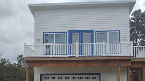 Une maison à deux étages avec un balcon, un garage et un terrain sablonneux où l’herbe pousse clairsemément.