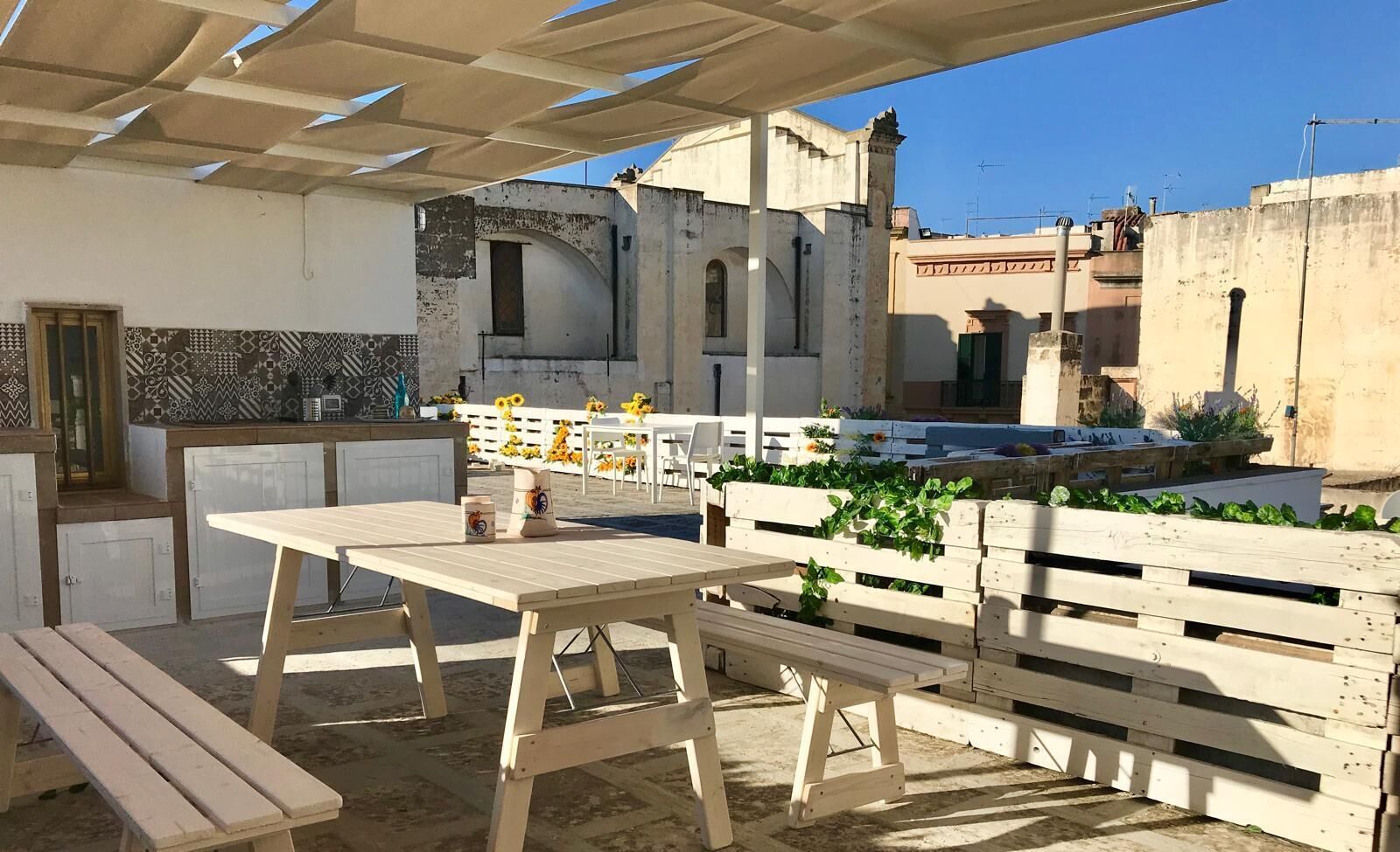 Een terras buiten met houten tafels en banken, een wit hek en uitzicht op een gebouw met een betegelde muur en gebogen ramen.